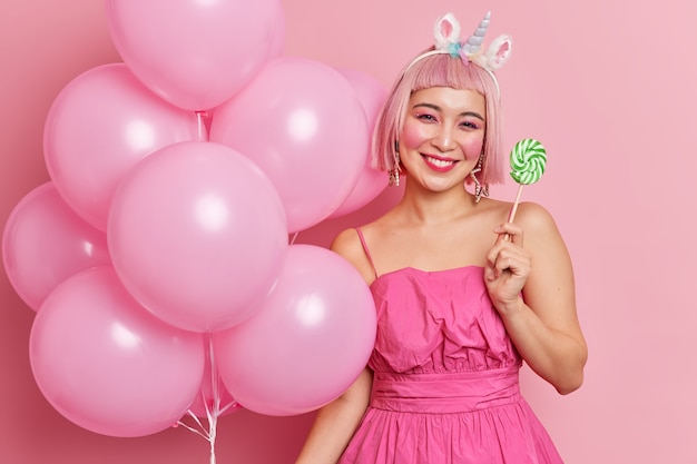 Positive Asian girl smiles gently has bright makeup wears dress holds delicious sweet candy bunch of inflated balloons enjoys holiday celebrates birthday being on party 