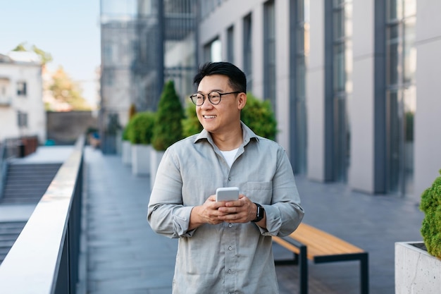 Positive asian middle aged male entrepreneur checking emails on smartphone while walking by business center