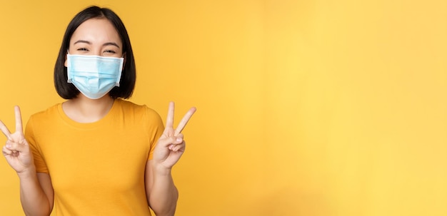 Positive asian woman smiling wearing medical face mask from covid during pandemic showing peace vsig...