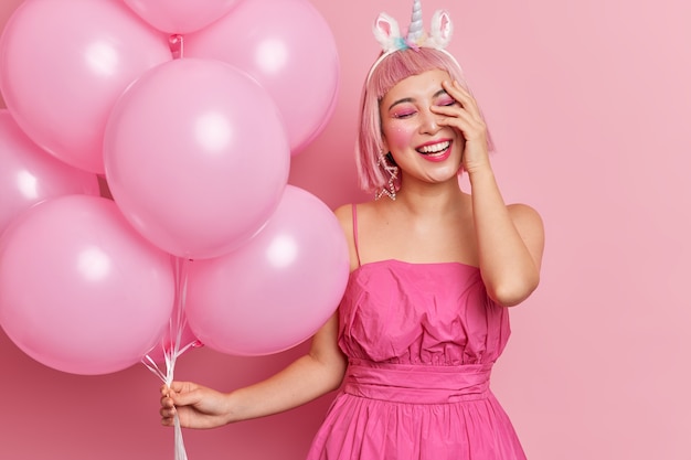 Positive Asian woman with pink hair giggles positively keeps hand on face has bright professional makeup wears dress has fun at party holds inflated helium balloons 