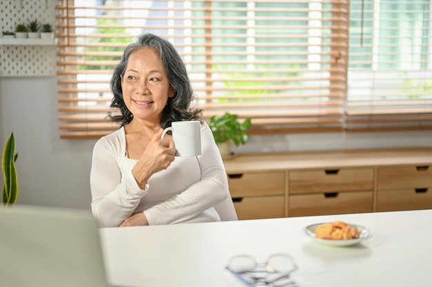 Positive Asianaged woman looking out the window daydreaming having her morning coffee