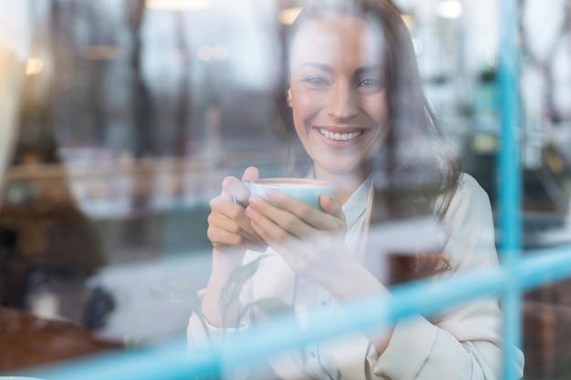 Positive atmosphere. Joyful merry positive woman sipping coffee while grinning and looking at the camera