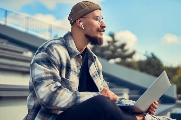 Positive bearded hipster guy sitting in outdoors and typing text on laptop g internet connection