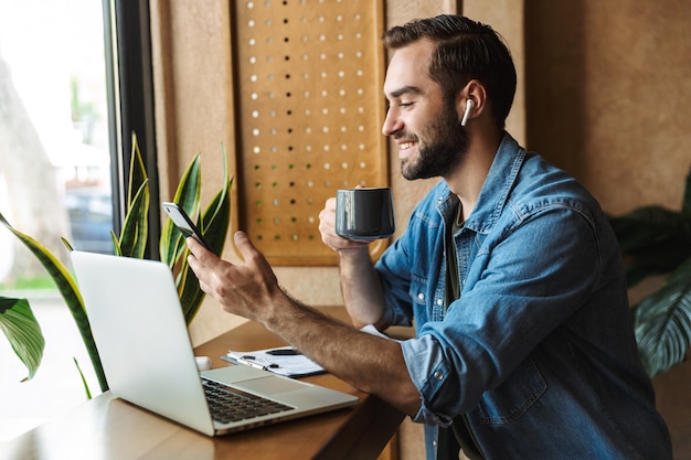 positive bearded man wearing denim shirt holding cup of tea and using smartphone while working in cafe indoors