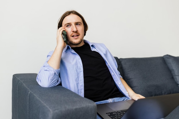 Positive bearded man with laptop talking on smartphone sitting on sofa in living room