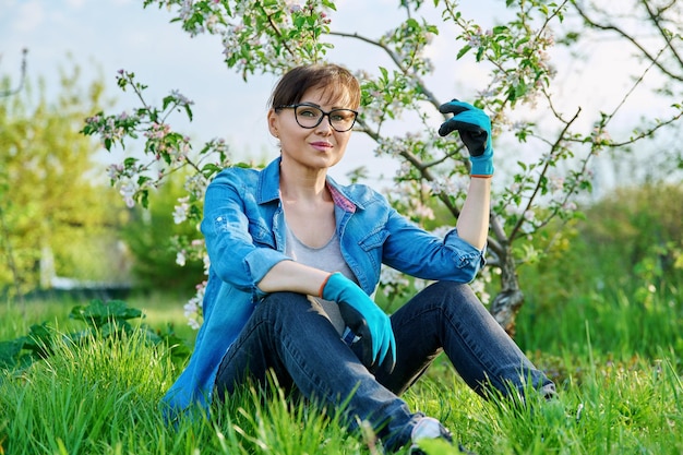 Positive beautiful mature female gardener sitting on the grass with blossoming apple tree