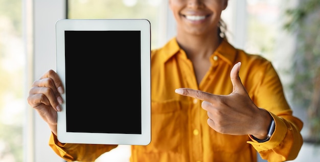 Positive black businesswoman pointing at digital tablet with blank screen in office interior cropped mockup