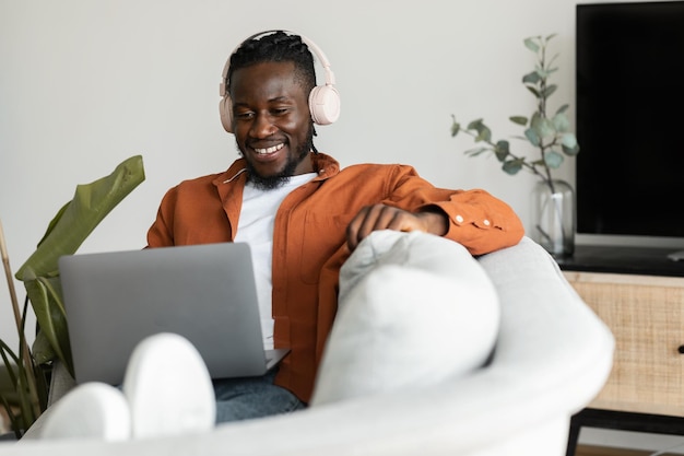 Positive black freelancer man in headphones working on laptop at home sitting on couch in living room free space