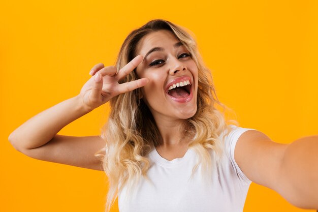 Positive blond woman smiling and taking selfie photo, isolated over yellow wall