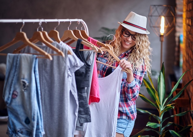 Positive blonde female in black eyeglasses chooses fashionable clothes on the coat rack.