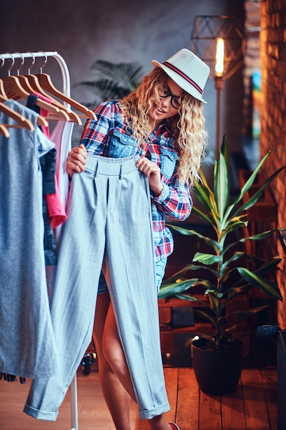 Positive blonde female in black eyeglasses chooses fashionable clothes on the coat rack.