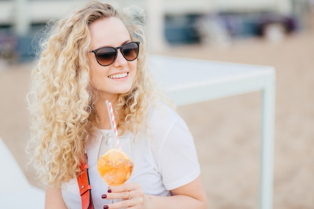 Positive blonde female with curly hair, wears sunglasses, looks with happy expression.