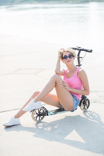 Positive blonde woman sitting at blue kick scooter