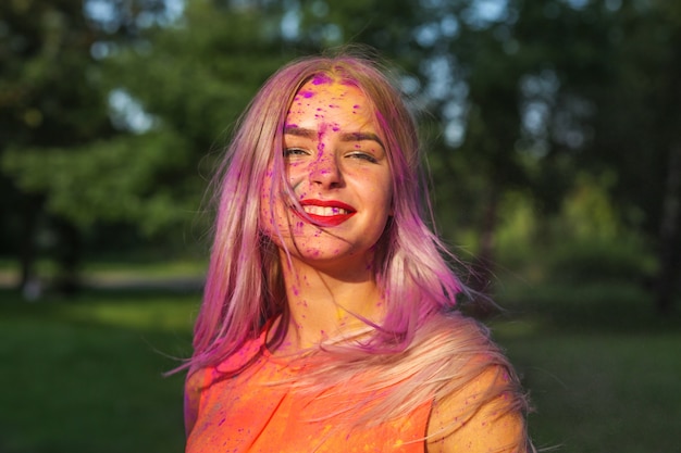 Positive blonde woman with long hair covered purple dry paint celebrating Holi festival