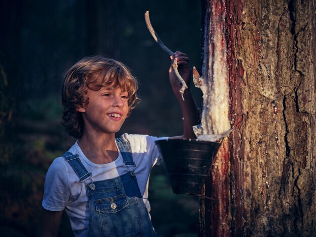 Positive boy with resin looking at wooden stick while standing near tree trunk with container in countryside on evening time