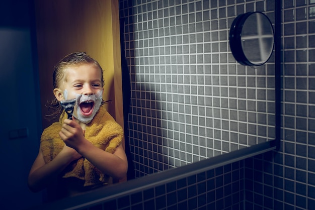 Positive boy with shaving foam on face and razor in hand looking at mirror while standing in bathroom during daily routine