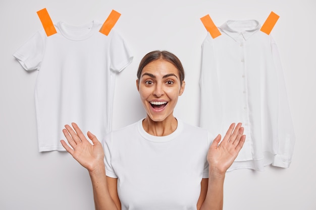 Photo positive brunette caucasian woman smiles broadly shows teeth raises palms poses against white wall with plastered clothes being in good mood