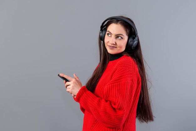 positive brunette in red sweater listens to music on smartphone in headphones isolated on gray