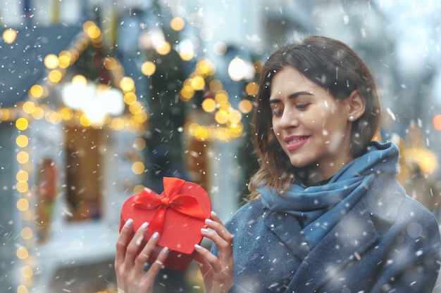 Positive brunette woman holding gift box near the christmas tree during the snowfall. Space for text