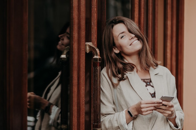 Positive brunette young woman with toothy smile wears smartwatch dressed in elegant jacket uses modern cell phone for online communication connected to wireless internet poses outdoors