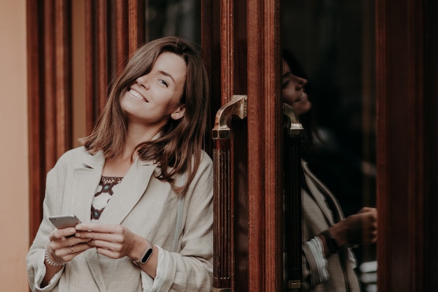 Positive brunette young woman with toothy smile, wears smartwatch, dressed in elegant jacket