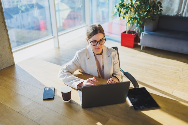 A positive business woman in a jacket is sitting at a table working on a laptop in a bright office A young European woman is sitting at a table Freelance and remote work Modern women's lifestyle