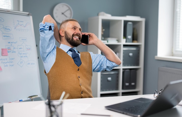 Positive businessman in stylish wear sitting at desk with laptop and talking on mobile Caucasian bearded male using modern gadgets for work at office