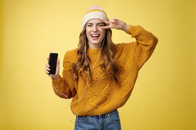 Positive carefree charming young girl show peace gesture wearing wired earphones showing smartphone screen promoting app cool brand new mobile phone, laughing carefree yellow background