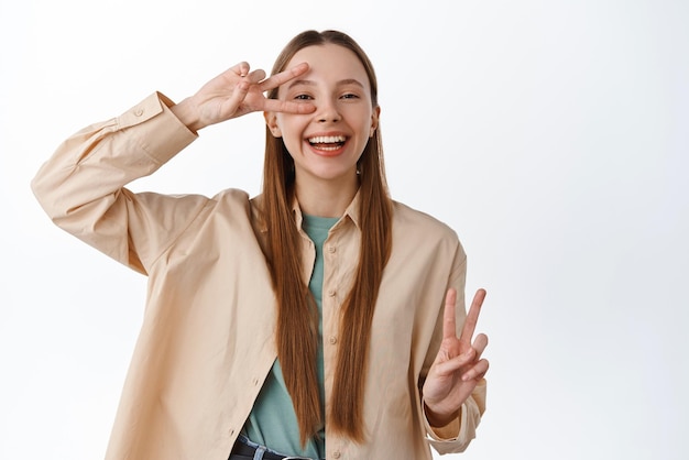 Positive caucasian girl student show peace sign and laughing happy making vsign gesture standing upbeat against white background