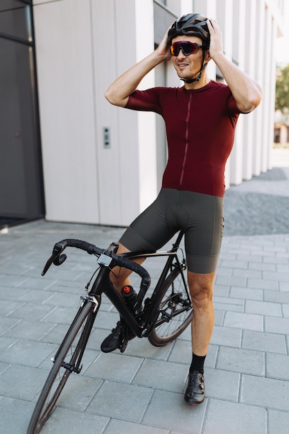 Positive caucasian man sitting on black bike and smiling while adjusting his safety helmet Caucasian cyclist enjoying favorite active hobby on fresh air