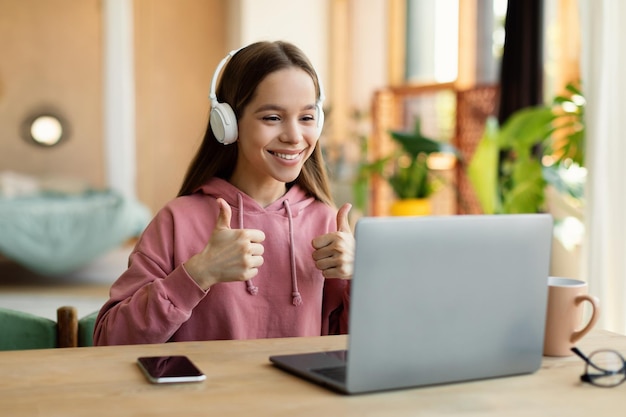 Positive caucasian teen girl in headphones showing thumb up while studying at home at table with