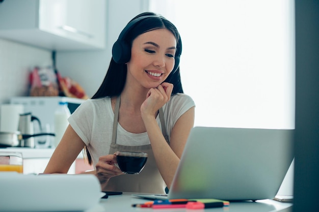 Positive Caucasian woman wearing headphones sitting at the table with a cup of coffee and looking at the screen of a modern laptop