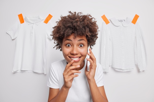 Positive cheerful woman has telephone conversation finds out amazing news smiles broadly dressed casually poses against white background with plastered clothes behind People and communication