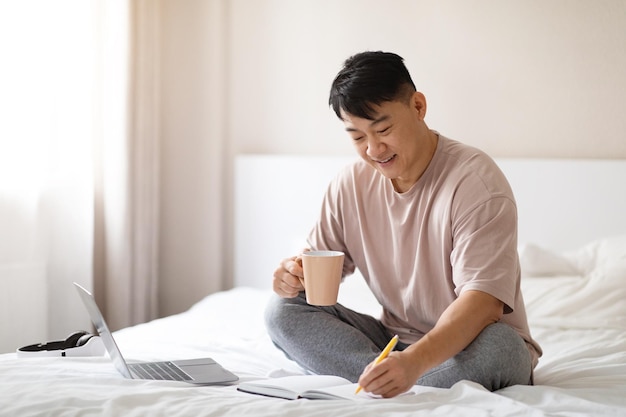 Positive chinese man planning vacation sitting on bed at home