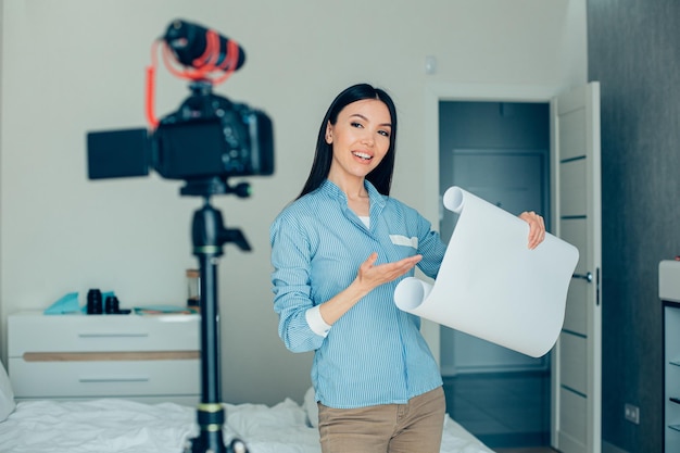 Positive confident young lady standing with a roll of architectural paper and smiling to the camera on tripod while demonstrating it