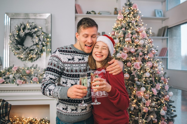 Positive couple stands together. Young man embrace woman. She keeps eyes closed and smiles. Each of them have glass of champaigne. People stand in decorated room.