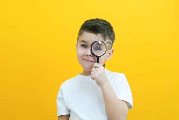 Positive curious schoolboy in casual clothes looks at the camera through a magnifying glass on a yellow background