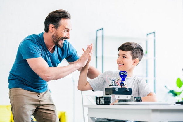 Positive delighted father giving high five to his son while finishing their engineering project on modern robot