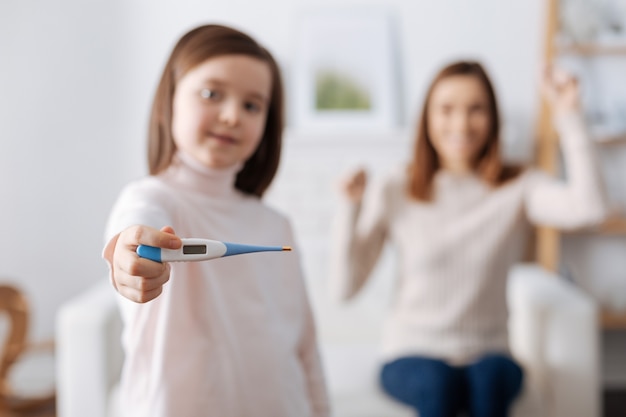 Positive delighted girl holding thermometer and feeling happy while her mother sitting in the background