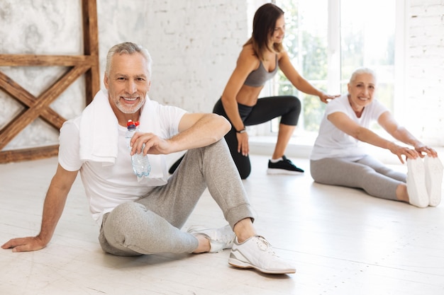 Positive delighted mature man keeping smile on his face and putting right hand on the floor while posing on camera