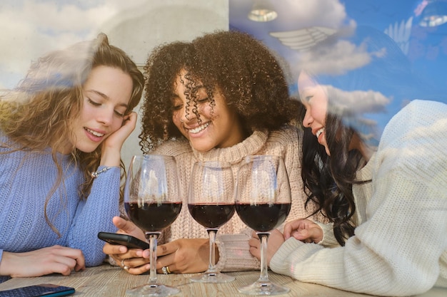 Photo positive diverse female friends in sweaters with glasses of red wine looking at smartphone screen