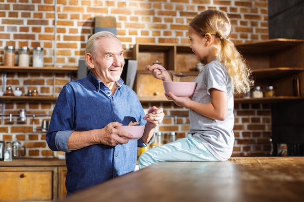 Positive elderly man eating cereal while enjoying time with his granddaughter