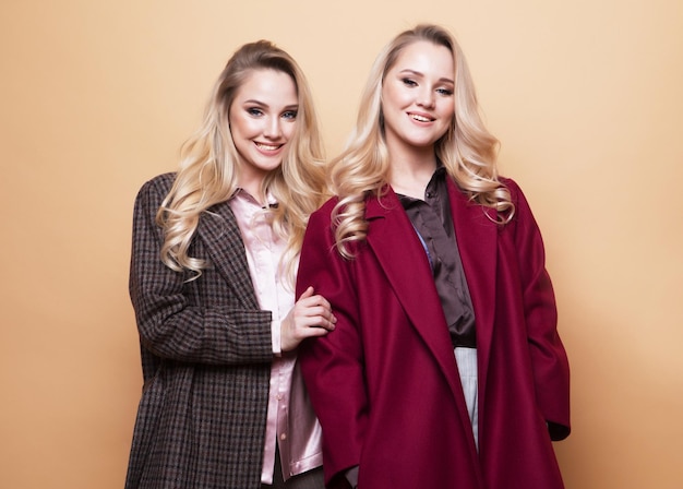 Positive fashion portrait of two girls best friends posing indoor on beige background wearing winter stylish coat