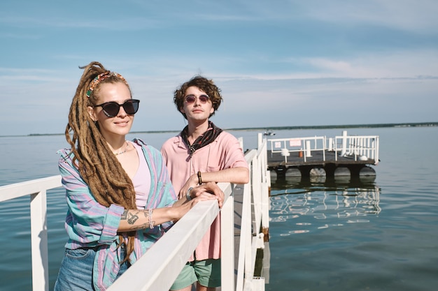 Photo positive fashionable young people standing on pier on lake posing for photo on sunny summer day