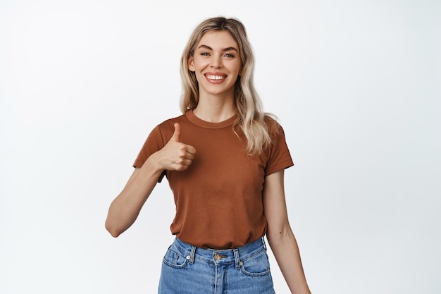 Positive feedback Smiling blond girl shows thumbs up to approve or praise something Young woman making compliment recommending product white background