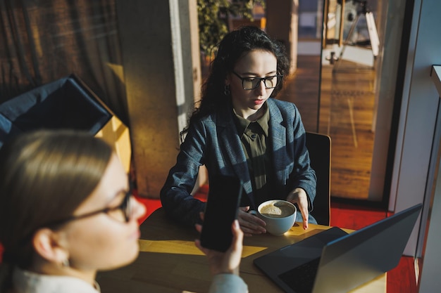 Positive female colleagues in casual clothes looking at netbook screen taking notes about project at table with coffee cups in office Students work Freelancers remote work Selective focus