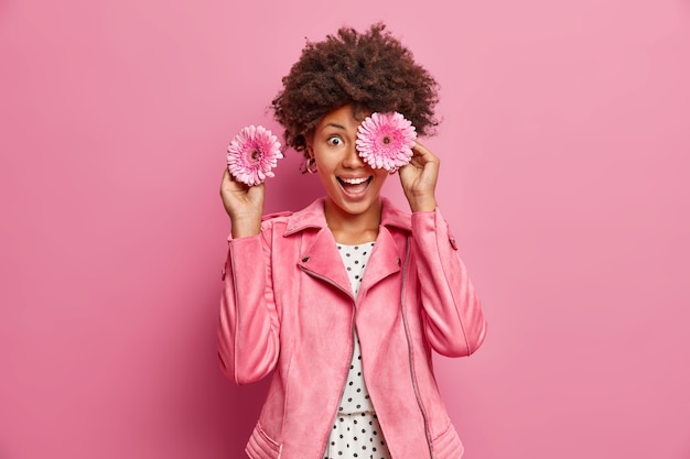 Positive female florist covers eyes with fragrant gerbera flowers makes bouquets for special occasions smiles broadly wears fashionable jacket isolated over pink wall