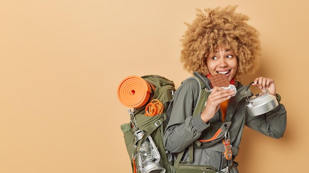 Positive female hiker has delicious snack bites bar of chocolate holds utensils and backpack with necessary equipment for travel looks somewhere gladfully isolated over brown background blank space