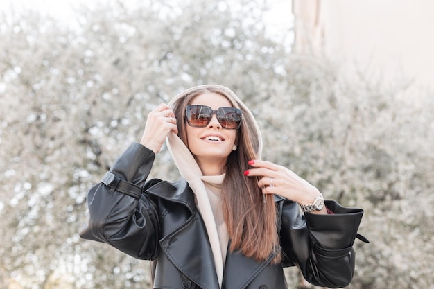 Positive female portrait of a beautiful stylish happy young woman with a smile with sunglasses in fashionable street clothes with a leather jacket and hoodie walking in the park and enjoying