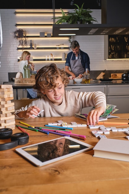 Positive focused boy with curly hair sitting at table and playing domino while parents making dinner in background